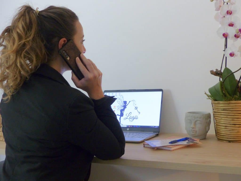 Megane assise de profil à son bureau, téléphone à l'oreille, devant son ordinater ouverte sur le logo de Logis Conciergerie
